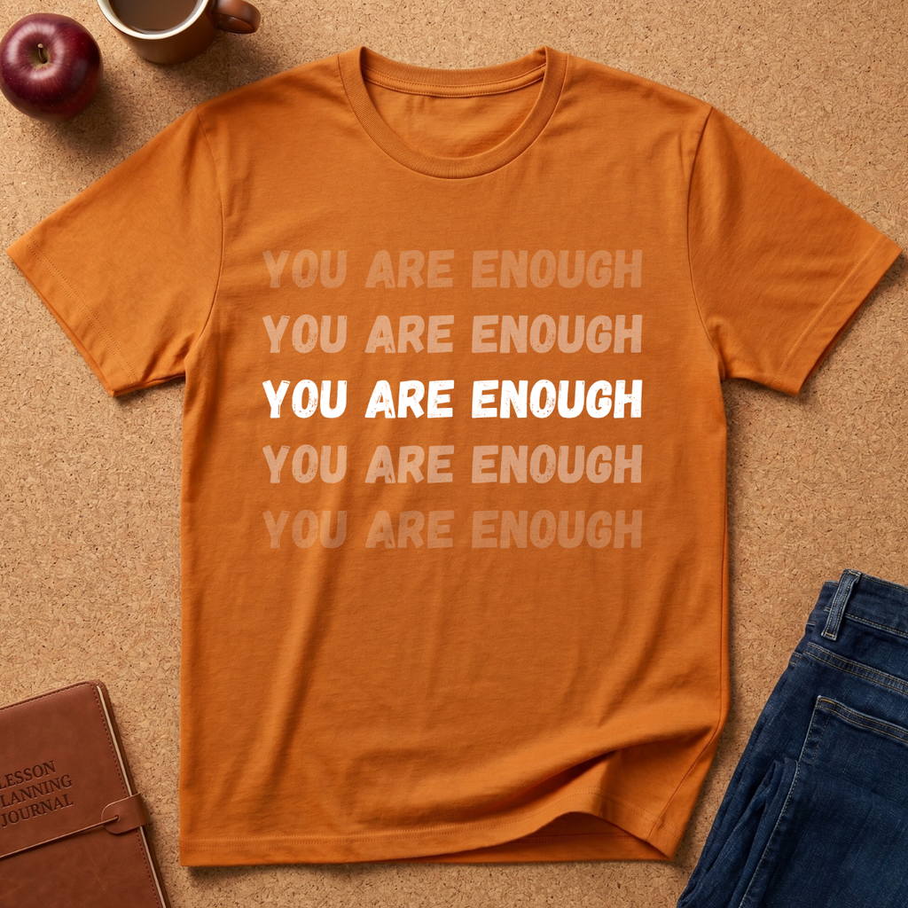 An orange short-sleeve t-shirt with the phrase 'YOU ARE ENOUGH' printed in white and faded orange text, presented in a flat-lay arrangement on a cork background with an apple, coffee mug, notebook, and jeans.