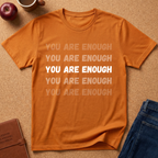 An orange short-sleeve t-shirt with the phrase 'YOU ARE ENOUGH' printed in white and faded orange text, presented in a flat-lay arrangement on a cork background with an apple, coffee mug, notebook, and jeans.