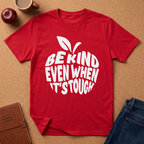 A red short-sleeve graphic T-shirt laid flat on a cork surface, featuring a white apple-shaped leaf graphic and the text 'BE KIND EVEN WHEN IT'S TOUGH.' A red apple, coffee mug, brown notebook, and blue jeans are placed around it as styling props.