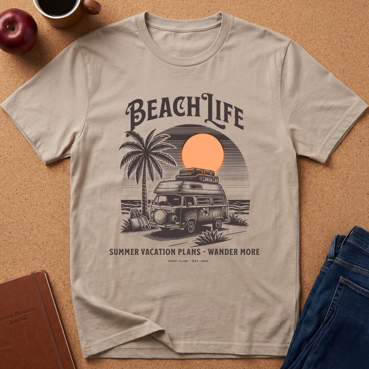 A light beige graphic t-shirt laid flat on a cork background, featuring a retro illustration of a camper van, palm tree, and sunset with the text 'BEACH LIFE' and 'SUMMER VACATION PLANS - WANDER MORE'. Surrounding props include an apple, coffee cup, notebook, and blue jeans.