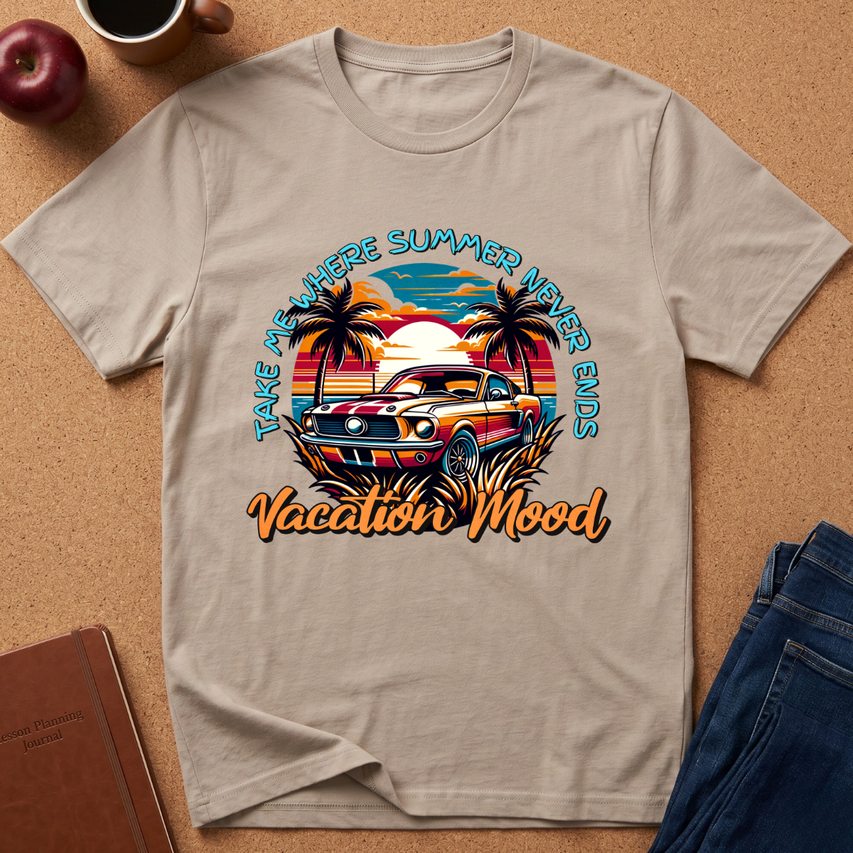 A beige graphic t-shirt laid flat on a cork background, featuring a retro-style illustration of a classic car at sunset with palm trees and the text 'TAKE ME HERE SUMMER VACATION MOOD' and 'Vacation Mood'. Props like an apple, coffee cup, notebook, and jeans are visible around the shirt.