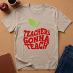 A light beige crewneck t-shirt with a graphic that reads 'TEACHERS GONNA TEACH' in red letters, topped with two green leaves, laid flat on a cork background with a coffee mug, apple, notebook, and jeans as props.