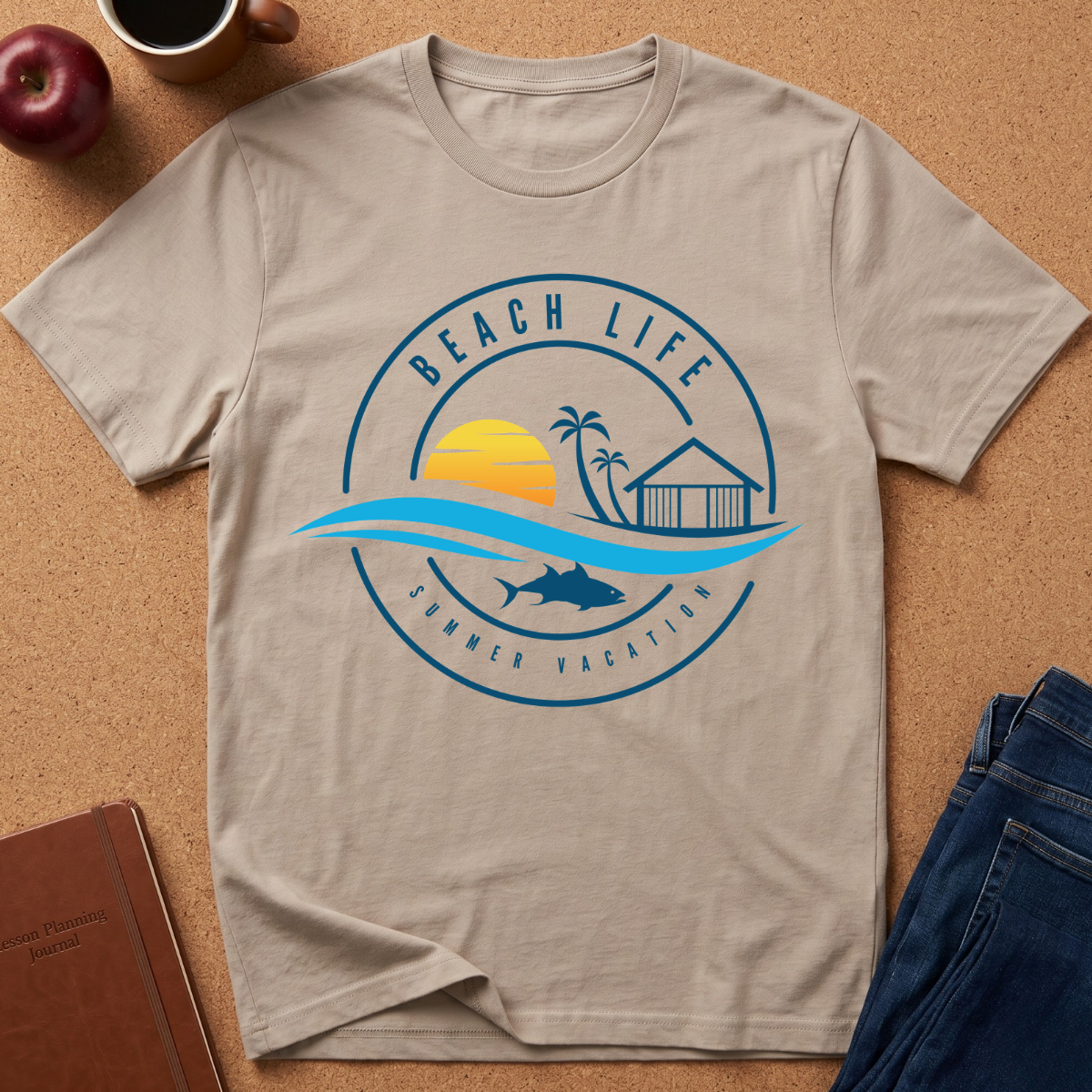 A light beige crewneck t-shirt with a blue and yellow 'Beach Life Summer Vacation' graphic print, laid flat on a cork background with lifestyle props including an apple, coffee cup, notebook, and jeans.