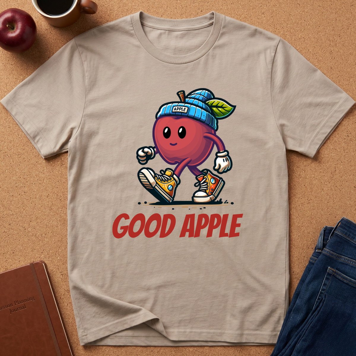 A beige graphic t-shirt laid flat on a cork background, featuring a cartoon red apple character wearing a blue knit beanie and sneakers with the text 'GOOD APPLE' below. Lifestyle props like an apple, coffee cup, notebook, and jeans are visible around the shirt.