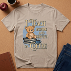 A beige graphic t-shirt laid flat on a cork background, featuring a cartoon teddy bear sitting on books with the text 'I TEACH INSPIRE AND SURVIVE ON COFFEE'. Props like an apple, coffee cup, notebook, and jeans are visible around the shirt.