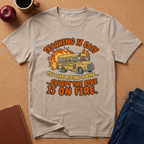 Beige t-shirt with a graphic design of a school bus and text on a brown surface with an apple and notebook. Teaching is easy It's like riding a bike.. except the Bike is on fire.