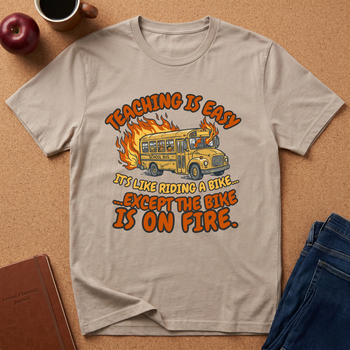 Beige t-shirt with a graphic design of a school bus and text on a brown surface with an apple and notebook. Teaching is easy It's like riding a bike.. except the Bike is on fire.