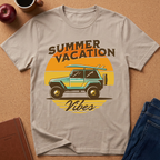 A light beige graphic t-shirt laid flat on a cork background, featuring a retro sunset design with a turquoise and yellow jeep carrying a surfboard, and the words 'SUMMER VACATION' and 'Vibes'.