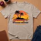 A light beige graphic t-shirt laid flat on a cork background, featuring a retro sunset design with palm trees and the text 'Dreaming of Summer Vacation NO MORE CLASSES'. Lifestyle props like an apple, coffee cup, notebook, and jeans are visible around the shirt.