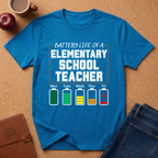Blue t-shirt with 'Battery Life of a Elementary School Teacher' graphic on a brown surface with a coffee cup, apple, and jeans.