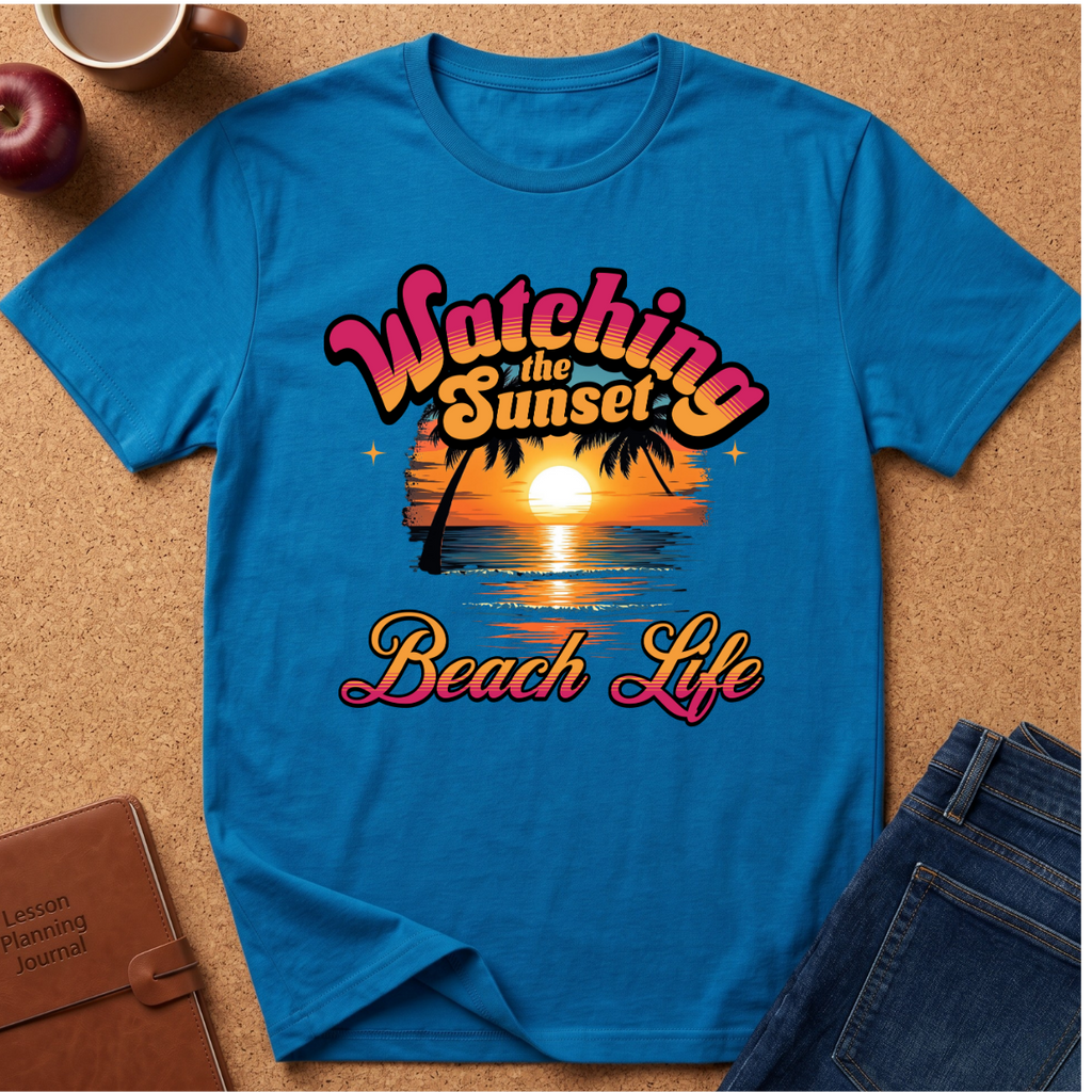 A bright blue short-sleeve graphic T-shirt laid flat on a cork surface, surrounded by styled props including an apple, a coffee mug, a journal, and jeans. The shirt features a retro sunset beach graphic with the text 'Watching the Sunset Beach Life' in pink, orange, yellow, and white.