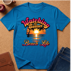 A bright blue short-sleeve graphic T-shirt laid flat on a cork surface, surrounded by styled props including an apple, a coffee mug, a journal, and jeans. The shirt features a retro sunset beach graphic with the text 'Watching the Sunset Beach Life' in pink, orange, yellow, and white.