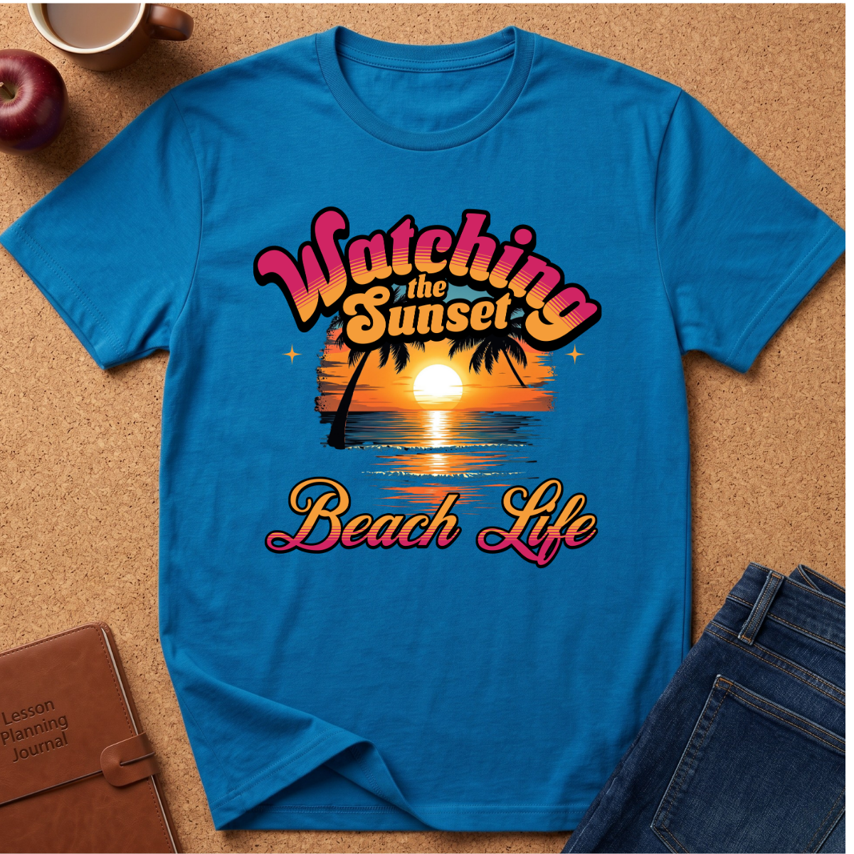 A bright blue short-sleeve graphic T-shirt laid flat on a cork surface, surrounded by styled props including an apple, a coffee mug, a journal, and jeans. The shirt features a retro sunset beach graphic with the text 'Watching the Sunset Beach Life' in pink, orange, yellow, and white.
