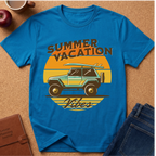 A bright blue graphic t-shirt laid flat on a cork surface, featuring a retro sunset design with a teal jeep, surfboard, and the words 'SUMMER VACATION Vibes.' Surrounding props include jeans, an apple, a coffee cup, and a journal.