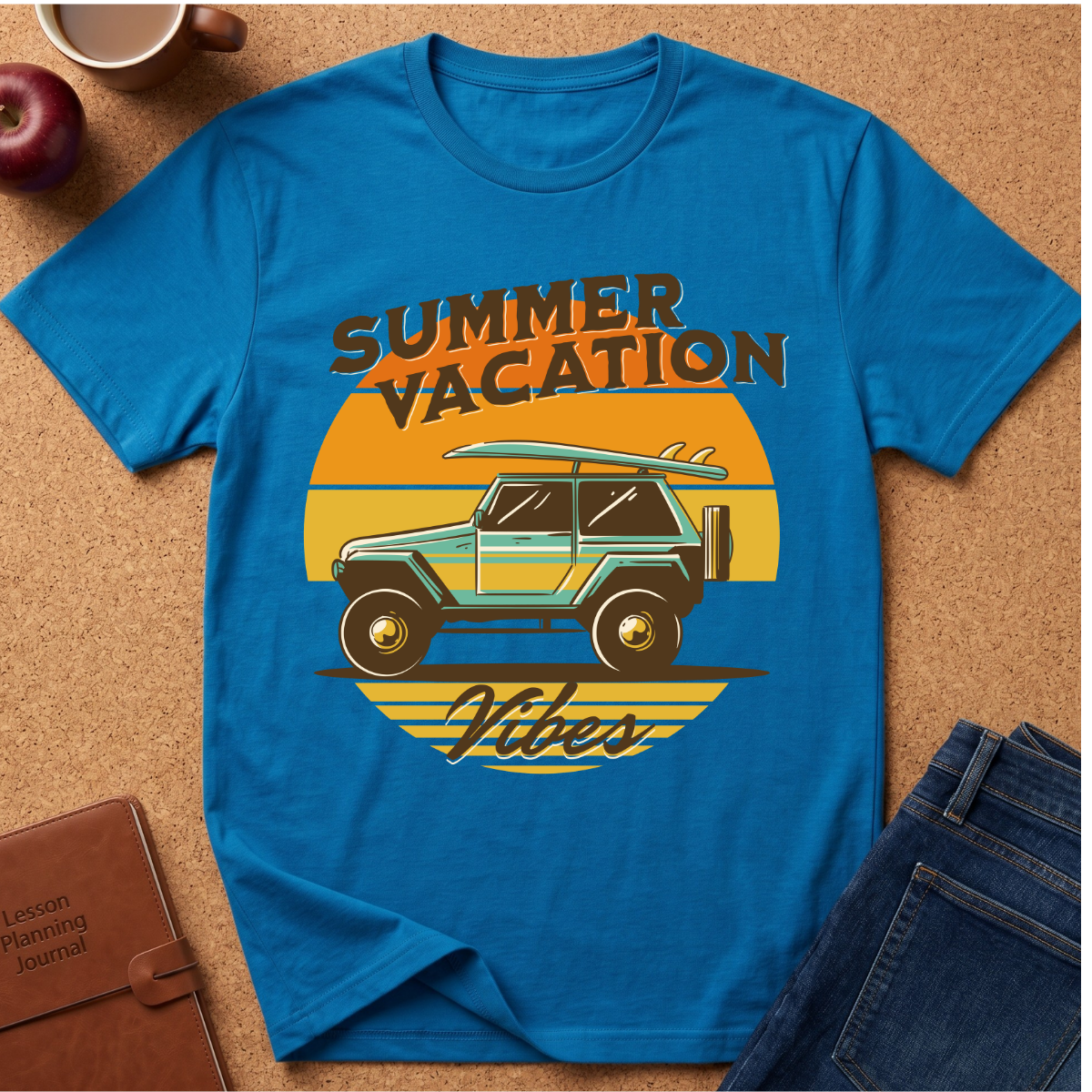 A bright blue graphic t-shirt laid flat on a cork surface, featuring a retro sunset design with a teal jeep, surfboard, and the words 'SUMMER VACATION Vibes.' Surrounding props include jeans, an apple, a coffee cup, and a journal.