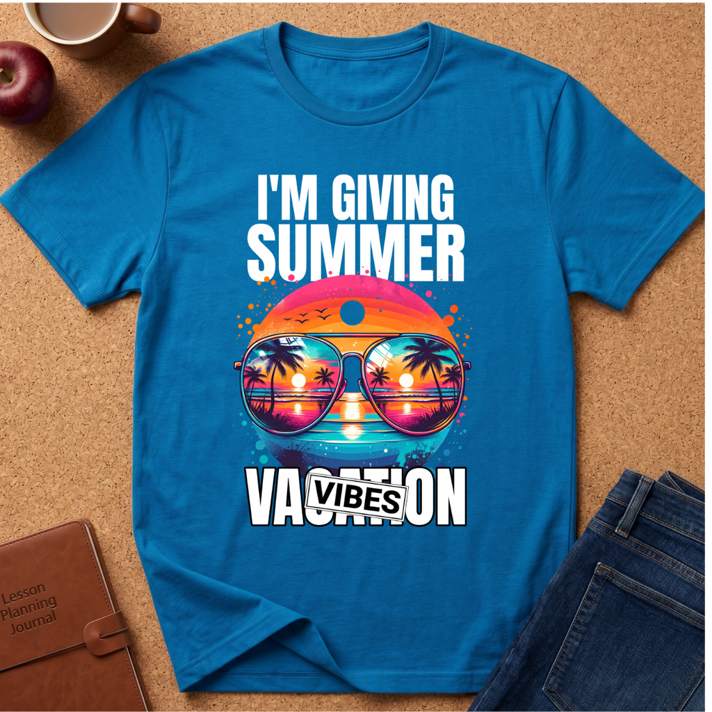 A bright blue graphic t-shirt laid flat on a cork surface, surrounded by lifestyle props including jeans, a brown journal, an apple, and a coffee cup. The t-shirt features a colorful graphic of sunglasses with a sunset and palm tree reflection, with the text 'I'M GIVING SUMMER VACATION VIBES'.