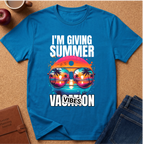 A bright blue graphic t-shirt laid flat on a cork surface, surrounded by lifestyle props including jeans, a brown journal, an apple, and a coffee cup. The t-shirt features a colorful graphic of sunglasses with a sunset and palm tree reflection, with the text 'I'M GIVING SUMMER VACATION VIBES'.
