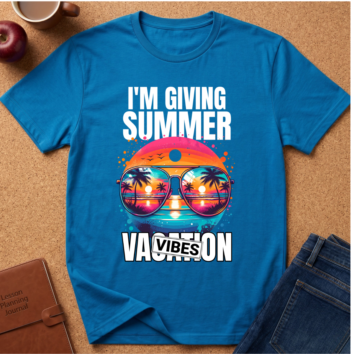 A bright blue graphic t-shirt laid flat on a cork surface, surrounded by lifestyle props including jeans, a brown journal, an apple, and a coffee cup. The t-shirt features a colorful graphic of sunglasses with a sunset and palm tree reflection, with the text 'I'M GIVING SUMMER VACATION VIBES'.