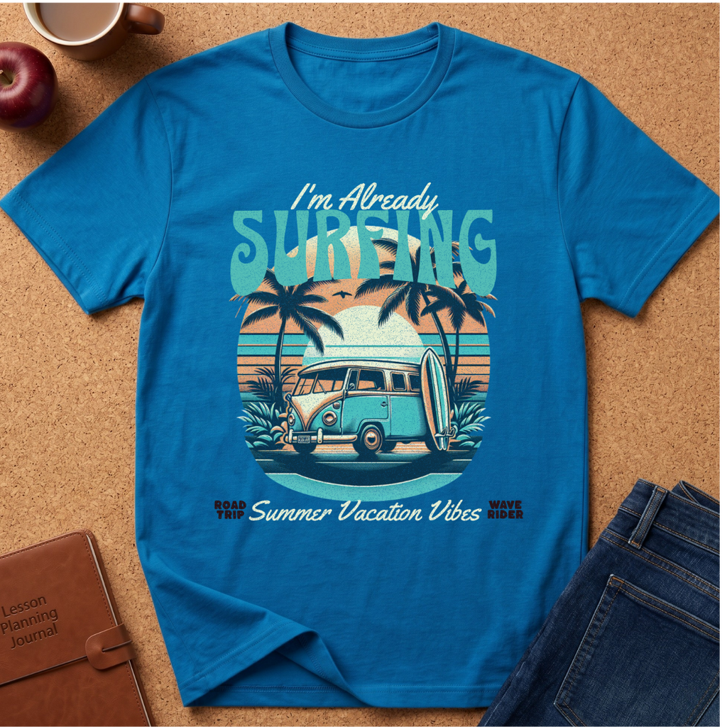 Saphire short-sleeve graphic T-shirt laid flat on a cork surface with styled props including an apple, coffee cup, notebook, and jeans. The shirt features a retro beach scene with palm trees, a van, surfboard, and the text 'I'm Already SURFING' and 'Summer Vacation Vibes'.