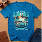 Saphire short-sleeve graphic T-shirt laid flat on a cork surface with styled props including an apple, coffee cup, notebook, and jeans. The shirt features a retro beach scene with palm trees, a van, surfboard, and the text 'I'm Already SURFING' and 'Summer Vacation Vibes'.