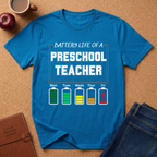 Blue t-shirt with 'Battery Life of a Preschool Teacher' graphic on a brown surface with accessories.