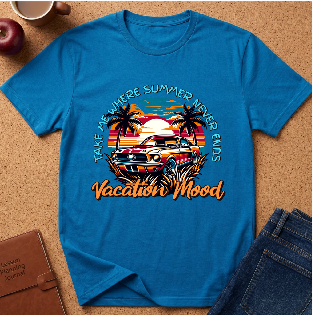 A bright blue graphic t-shirt laid flat on a cork background, featuring a retro-style tropical car scene with palm trees, a sunset, and the text 'TAKE ME WHERE SUMMER NEVER ENDS Vacation Mood'. Styled flat-lay props include jeans, a notebook, an apple, and a coffee cup.
