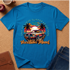 A bright blue graphic t-shirt laid flat on a cork background, featuring a retro-style tropical car scene with palm trees, a sunset, and the text 'TAKE ME WHERE SUMMER NEVER ENDS Vacation Mood'. Styled flat-lay props include jeans, a notebook, an apple, and a coffee cup.