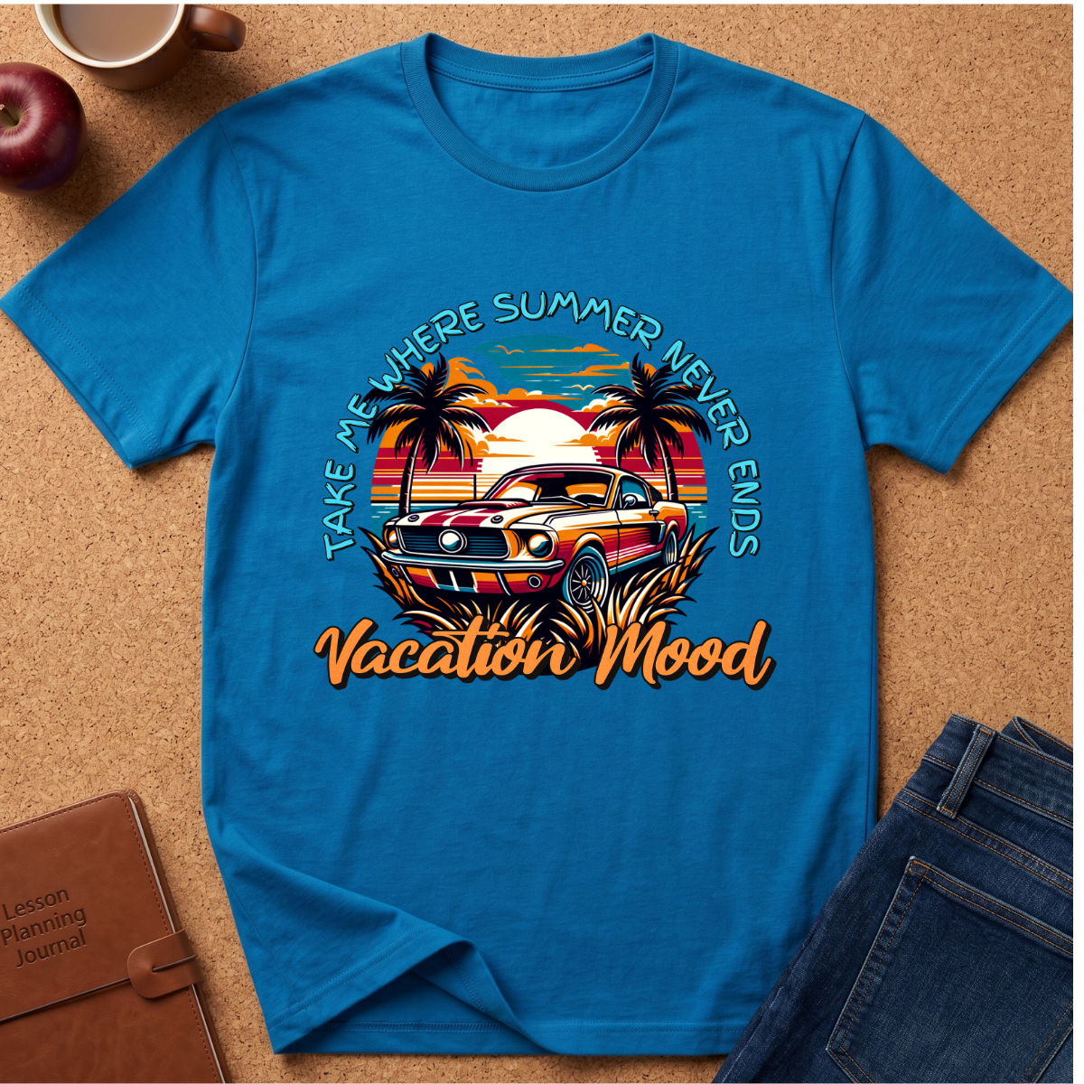 A bright blue graphic t-shirt laid flat on a cork background, featuring a retro-style tropical car scene with palm trees, a sunset, and the text 'TAKE ME WHERE SUMMER NEVER ENDS Vacation Mood'. Styled flat-lay props include jeans, a notebook, an apple, and a coffee cup.