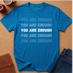 A bright blue graphic t-shirt laid flat on a cork background, featuring the phrase 'YOU ARE ENOUGH' repeated in light blue, white, and dark blue text. Styled flat-lay props include a brown notebook, a red apple, a coffee mug, and blue jeans.