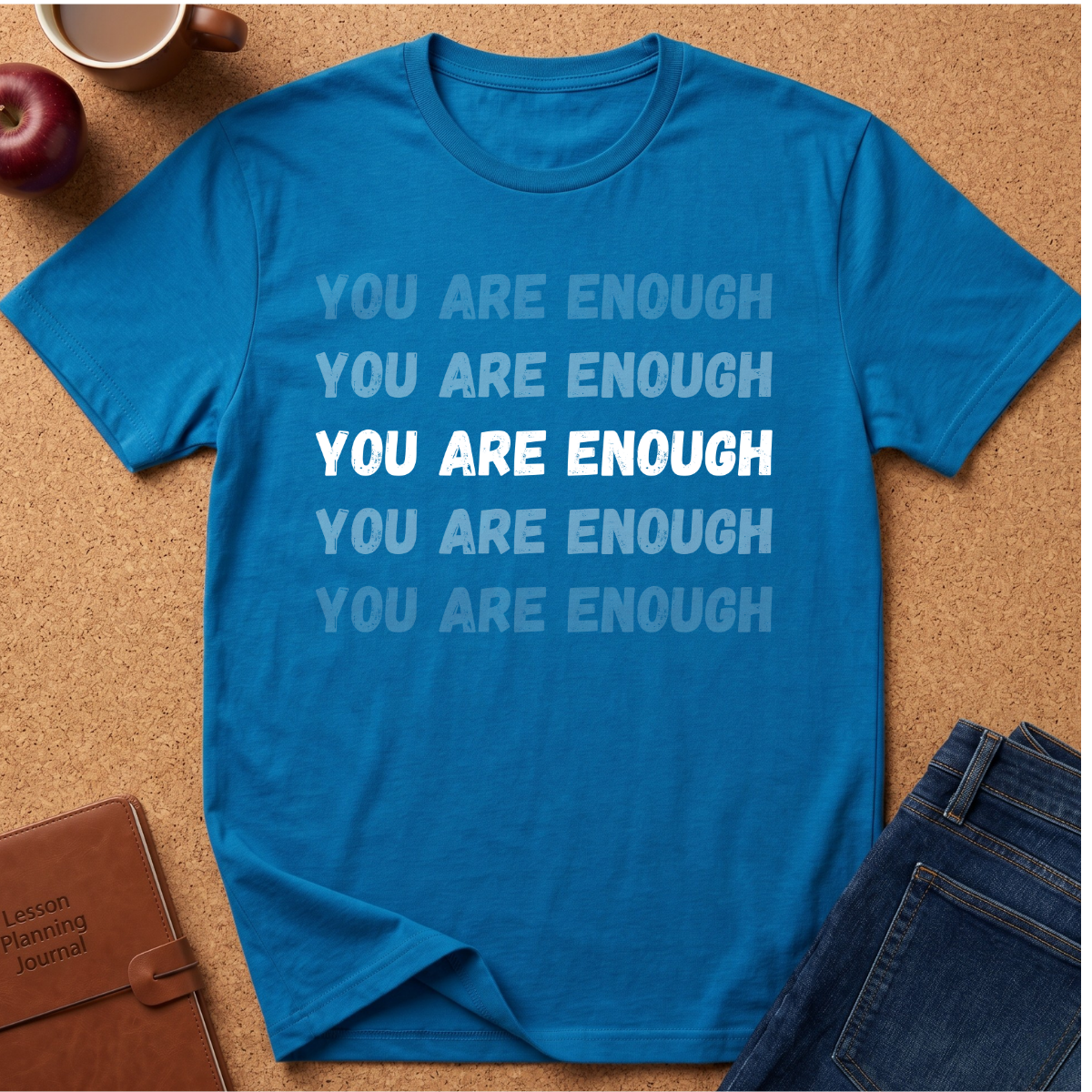 A bright blue graphic t-shirt laid flat on a cork background, featuring the phrase 'YOU ARE ENOUGH' repeated in light blue, white, and dark blue text. Styled flat-lay props include a brown notebook, a red apple, a coffee mug, and blue jeans.