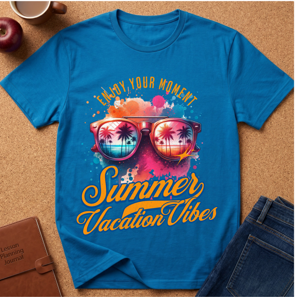 A bright blue graphic T-shirt laid flat on a cork surface, surrounded by a coffee cup, apple, notebook, and jeans. The shirt features a colorful retro design with sunglasses reflecting a tropical beach scene and the text 'Enjoy Your Moment Summer Vacation Vibes'.