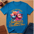 A bright blue graphic T-shirt laid flat on a cork surface, surrounded by a coffee cup, apple, notebook, and jeans. The shirt features a colorful retro design with sunglasses reflecting a tropical beach scene and the text 'Enjoy Your Moment Summer Vacation Vibes'.