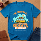 A bright blue graphic t-shirt laid flat on a cork surface, surrounded by lifestyle props including a brown leather journal, an apple, a coffee cup, and folded blue jeans. The t-shirt features a retro-style graphic of a yellow and white van with a surfboard on its roof, set against a light blue cloud background with the text 'I AM ALREADY ON MY sUMMER VACATION. #SEEYOUINSEPTEMBER