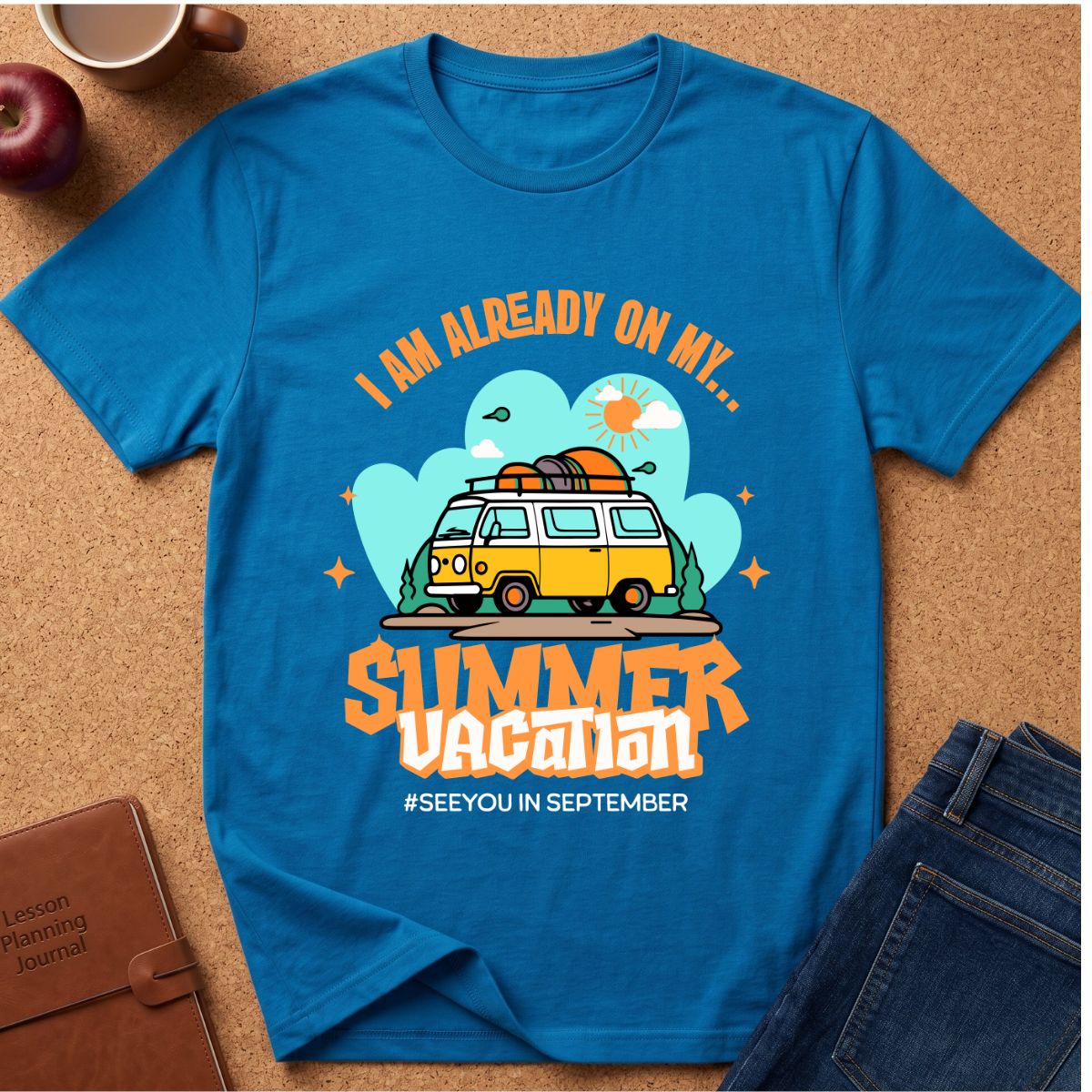A bright blue graphic t-shirt laid flat on a cork surface, surrounded by lifestyle props including a brown leather journal, an apple, a coffee cup, and folded blue jeans. The t-shirt features a retro-style graphic of a yellow and white van with a surfboard on its roof, set against a light blue cloud background with the text 'I AM ALREADY ON MY sUMMER VACATION. #SEEYOUINSEPTEMBER