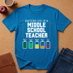 Blue t-shirt with 'Battery Life of a Middle School Teacher' graphic on a brown surface with a coffee cup, apple, and jeans.
