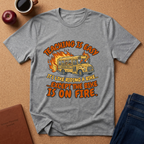 Gray t-shirt with a graphic of a school bus on fire and text, placed on a brown surface with an apple and coffee cup. Teaching is easy It's like riding a bike.. except the Bike is on fire.