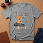 A heather gray graphic t-shirt laid flat on a cork background, featuring a design of a teddy bear sitting on books with the text 'I TEACH INSPIRE AND SURVIVE ON COFFEE'. The shirt is styled with a coffee mug, an apple, a notebook, and jeans.