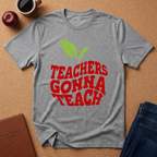 A heather gray short-sleeve t-shirt with a graphic of a green apple and the text 'TEACHERS GONNA TEACH' in red, laid flat on a cork background with a coffee cup, apple, and jeans nearby.