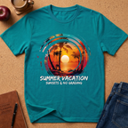 A teal short-sleeve graphic T-shirt laid flat on a cork surface, surrounded by styled props including a notebook, pen, apple, coffee cup, and folded jeans. The shirt features a circular tropical sunset print with palm trees and the text 'SUMMER VACATION SUNSETS & NO GRADING.