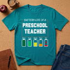 Teal t-shirt with 'Battery Life of a Preschool Teacher' graphic on a cork background with apple, coffee, and jeans.