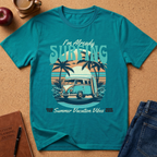 A teal short-sleeve graphic T-shirt laid flat on a cork surface with styled props including an apple, coffee cup, notebook, and jeans. The shirt features a retro beach scene with palm trees, a van, surfboard, and the text 'I'm Already SURFING' and 'Summer Vacation Vibes'.