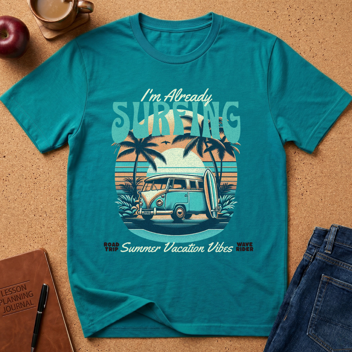 A teal short-sleeve graphic T-shirt laid flat on a cork surface with styled props including an apple, coffee cup, notebook, and jeans. The shirt features a retro beach scene with palm trees, a van, surfboard, and the text 'I'm Already SURFING' and 'Summer Vacation Vibes'.