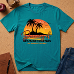 A teal short-sleeve graphic T-shirt laid flat on a cork-like surface, featuring a retro sunset beach scene with palm trees, birds, and the text 'Dreaming of Summer Vacation No More Classes.' Surrounding props include an apple, a coffee cup, a notebook, and blue jeans.
