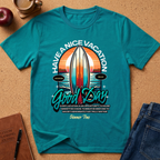 A teal short-sleeve graphic T-shirt laid flat on a cork surface, surrounded by lifestyle props including an apple, a notebook, a pen, and folded blue jeans. The shirt features a large retro-style surfboard and sunset graphic with the text 'HAVE A NICE VACATION Good Day' and smaller motivational text below.