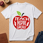 A white short-sleeve T-shirt laid flat on a cork background, featuring a large red apple graphic with the words 'TEACH INSPIRE GROW' in white and a green leaf at the top. Surrounding props include an apple, a coffee cup, a journal, and jeans.