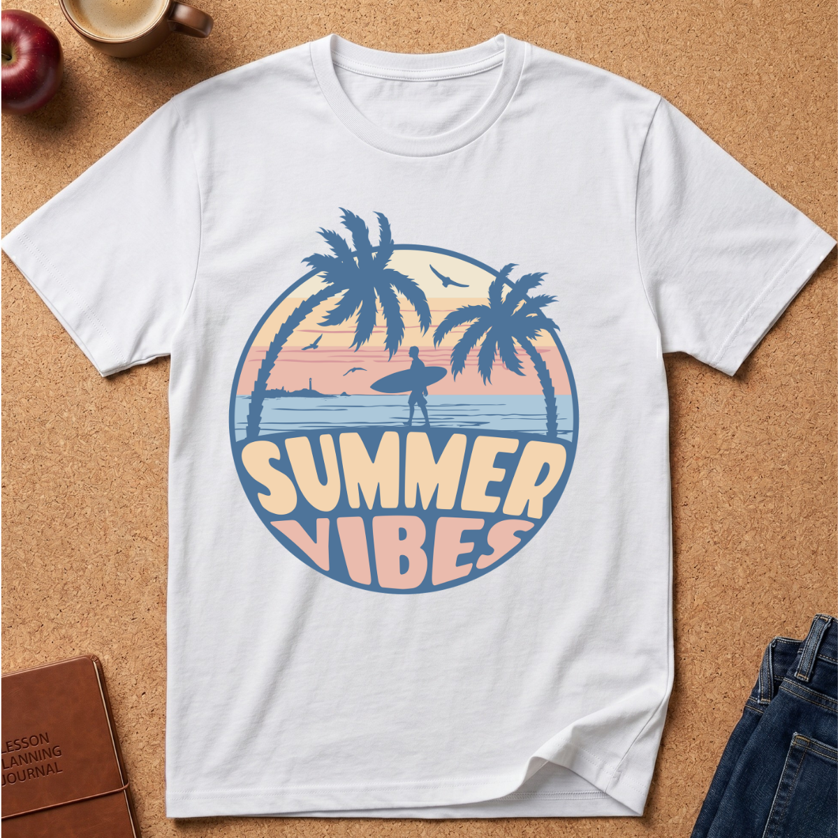 A white short-sleeve crewneck t-shirt laid flat on a cork board background, featuring a retro circular graphic with palm trees, a surfer silhouette, and the words 'SUMMER VIBES' in pastel colors. Styled flat-lay props include a brown journal, a blue denim jacket, and a red apple.