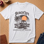 A white short-sleeve graphic T-shirt laid flat on a cork surface, featuring a retro beach-themed print with the words “BEACH LIFE,” a palm tree, a vintage camper van, a sunset circle, and smaller text reading “SUMMER VACATION PLANS - WANDER MORE.” Partial props including an apple, a coffee cup, a book, and jeans are visible around the shirt.