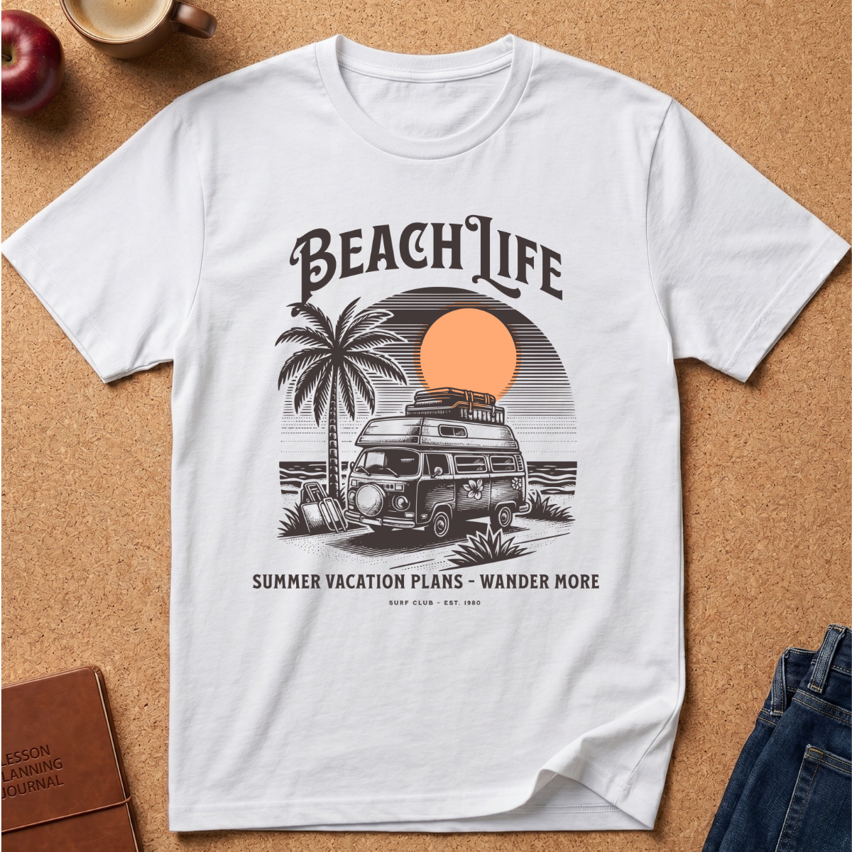 A white short-sleeve graphic T-shirt laid flat on a cork surface, featuring a retro beach-themed print with the words “BEACH LIFE,” a palm tree, a vintage camper van, a sunset circle, and smaller text reading “SUMMER VACATION PLANS - WANDER MORE.” Partial props including an apple, a coffee cup, a book, and jeans are visible around the shirt.