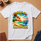 A white short-sleeve crewneck t-shirt laid flat on a cork background, featuring a colorful graphic print of a tropical beach scene with palm trees and the text 'SCHOOL'S OUT FOR SUMMER' in a rainbow gradient.