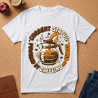 White graphic t-shirt says "Current Status, Powered by Caffeine and Chaos" with coffee-themed graphic and text on a brown surface with coffee and an apple.